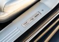 Close-up of a metal door sill plate featuring Aston Martin logos and "DBS Superleggera" branding beside a white car seat and black trim.