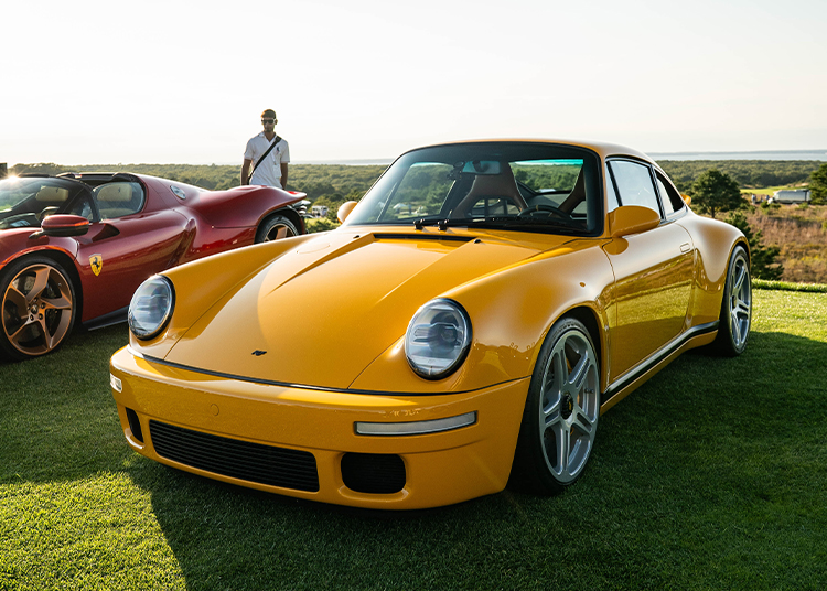 A yellow sports car, showcasing true automotive art, is parked on grass next to a red car, with a person standing in the background under a clear sky.