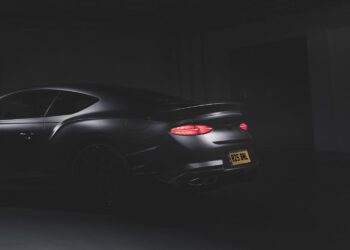 A matte black Bentley sports car is shown from the rear in a dimly lit setting, its taillights glowing and a yellow license plate visible—a rare car from a 100-year-old name.