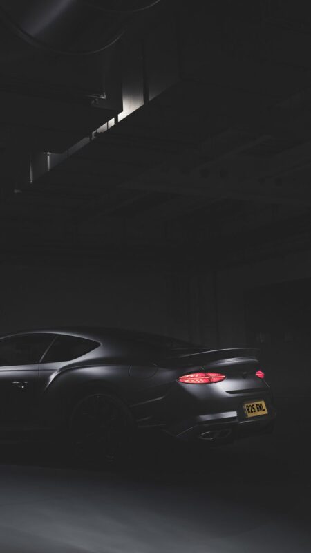 A matte black Bentley sports car with illuminated tail lights, a rare car from the 100-year-old name, is parked in a dimly lit industrial garage.