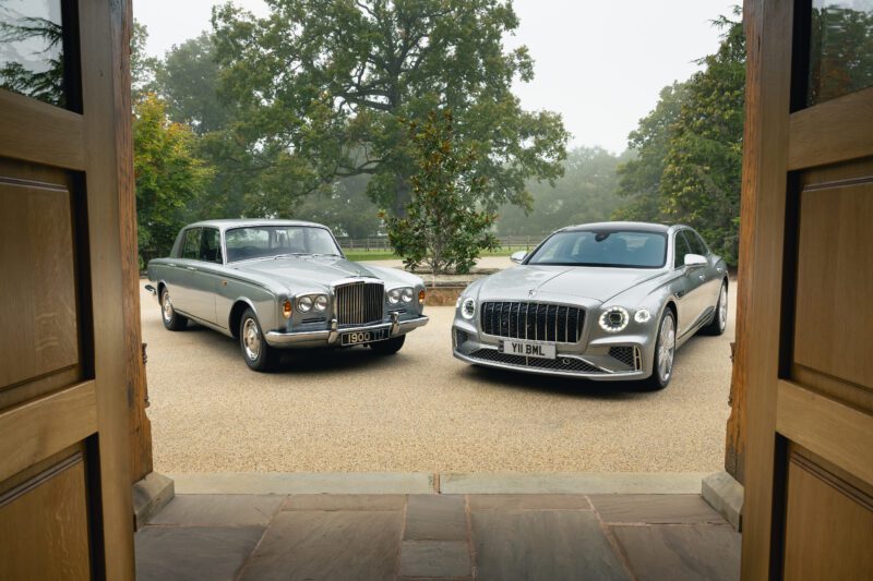 A classic silver Rolls-Royce and a modern silver Bentley sedan, embodying Legendary Sedans, are parked side by side on a driveway, framed by open wooden doors and surrounded by trees.