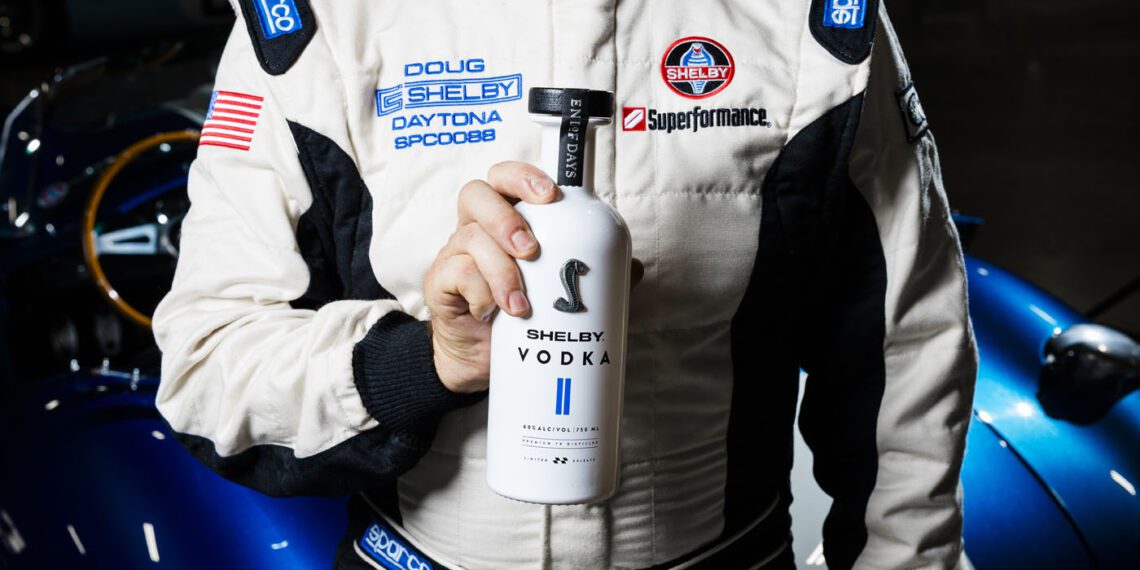 A person in a racing suit holds a bottle of Shelby Vodka, capturing the spirit of a legend in front of a blue sports car.