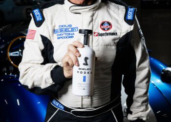A person in a racing suit holds a bottle of Shelby Vodka, capturing the spirit of a legend in front of a blue sports car.