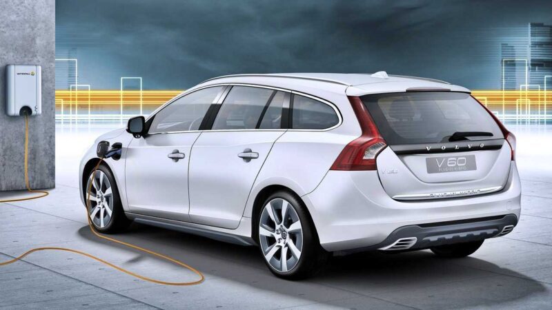 A white Volvo V60 is parked and charging at a wall-mounted electric vehicle charger, its cable connected to the car’s front left side—a testament to Volvo’s global plug-in hybrid sales crossing one million.