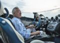 An older man wearing glasses sits in the driver's seat of a Pagani hypercar with a blue and white interior at Auto e Moto d'Epoca 2025, looking out the window.
