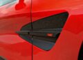 Close-up of a red car’s side vent featuring a black trim with “Aston Martin V12” badge. Review: 2025 Aston Martin Vanquish Coupe.