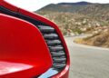 Close-up of the rear taillight and body of a red sports car, likely the 2025 Aston Martin Vanquish Coupe, parked on a winding mountain road with hills in the background.
