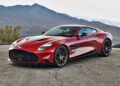 A red 2025 Aston Martin Vanquish Coupe is parked on a paved surface, with mountains and a cloudy sky in the background. Review: This sleek sports car blends elegance and power against a stunning natural backdrop.