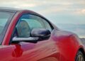 Close-up of the driver's side door and mirror of a red sports car, with a scenic, blurred background of mountains and sky—perfect for a Review: 2025 Aston Martin Vanquish Coupe.
