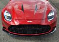 Front view of a red 2025 Aston Martin Vanquish Coupe sports car showing its grille, headlights, hood vents, and emblem, parked on a paved surface. Review: Stunning design and performance highlights the latest Vanquish.