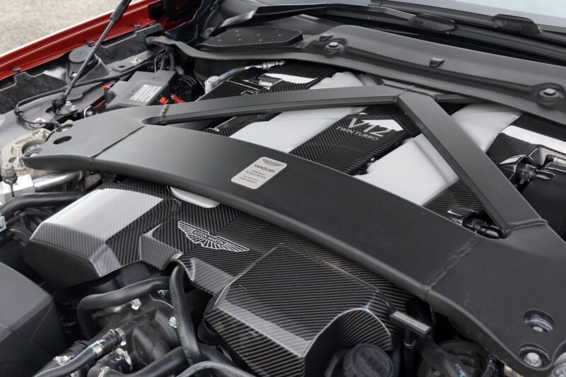 Close-up of a V12 engine bay with visible carbon fiber components and the Aston Martin logo, highlighting detailed engine parts and structural bracing in the Review: 2025 Aston Martin Vanquish Coupe.