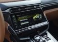 Close-up of a car’s infotainment screen displaying drive mode options, surrounded by dashboard controls, air vents, and tan interior accents in the Review: 2025 Aston Martin Vanquish Coupe.