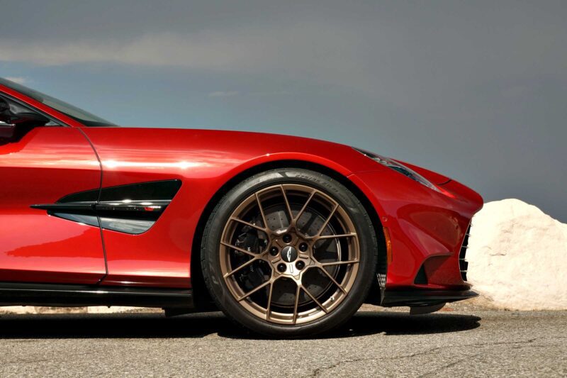 Close-up of the front side of a red Review: 2025 Aston Martin Vanquish Coupe with bronze alloy wheels parked on pavement, featuring a white rock and gray sky in the background.