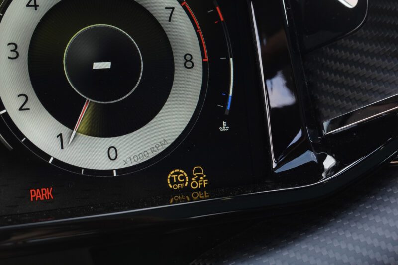 Close-up of a car dashboard in the Review: 2025 Aston Martin Vanquish Coupe, showing part of the tachometer, red "PARK" indicator, and yellow warning lights for traction control and stability control system.