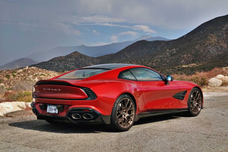 A red 2025 Aston Martin Vanquish Coupe is parked on a mountain road, with rolling hills and a cloudy sky in the background. Review: This stunning sports car blends luxury and performance in an unforgettable setting.