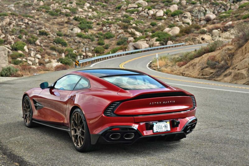 A red 2025 Aston Martin Vanquish Coupe is parked on the side of a winding mountain road with rocky hills in the background. Review: experience elegance and performance in every curve.