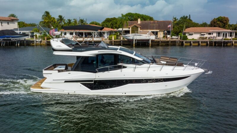 A modern white yacht glides through a calm waterfront channel, offering a true maritime experience as it passes houses and docks lined with palm trees beneath a cloudy sky.
