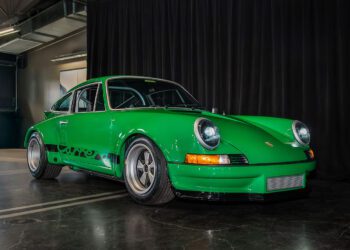 A green 2025 Porsche, a National Winner in Porsche Classic Restoration, is parked indoors on a polished concrete floor in front of a black curtain.