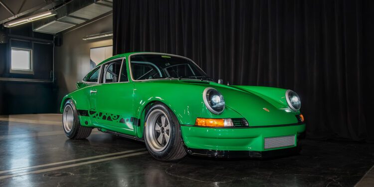 A green 2025 Porsche, a National Winner in Porsche Classic Restoration, is parked indoors on a polished concrete floor in front of a black curtain.