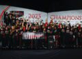 A large Porsche racing team celebrates on stage in front of a “2023 Champions” banner, holding a sign that reads “2023 IMSA WeatherTech SportsCar Championship,” looking ahead to 2025 with excitement.