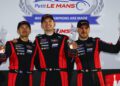 Three race car drivers in matching suits hold trophies and smile on the podium at the 2025 Petit Le Mans, part of the IMSA WeatherTech SportsCar Championship. A backdrop with event and Porsche branding is visible behind them.