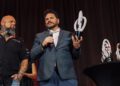 Two men stand on stage; one holds a microphone and a modern white trophy, while another similar trophy sits on a nearby table.
