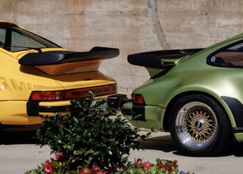 A yellow and a green Porsche 911 Turbo parked side by side, viewed from the rear, at Luftgekühlt 11 in Durham NC, with a concrete wall in the background and plants in the foreground.