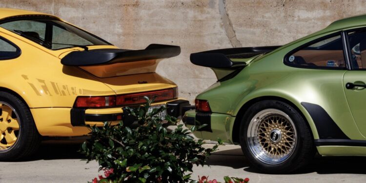 A yellow and a green Porsche 911 Turbo parked side by side, viewed from the rear, at Luftgekühlt 11 in Durham NC, with a concrete wall in the background and plants in the foreground.