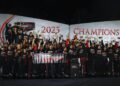 A Porsche racing team poses together at night celebrating their 2023 IMSA WeatherTech SportsCar championship win, holding a banner and trophies in front of a backdrop that reads "2023 Champions.