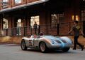 A vintage silver Porsche 993 with the number 86 drives on a street in Durham NC next to a brick building, as a person in a "Crew" shirt walks nearby.