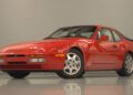 A red Porsche 944 sports car, featured in the 2025 Porsche Classic Restoration Challenge, is parked indoors on a glossy floor, facing left, with its headlights closed and windows up.