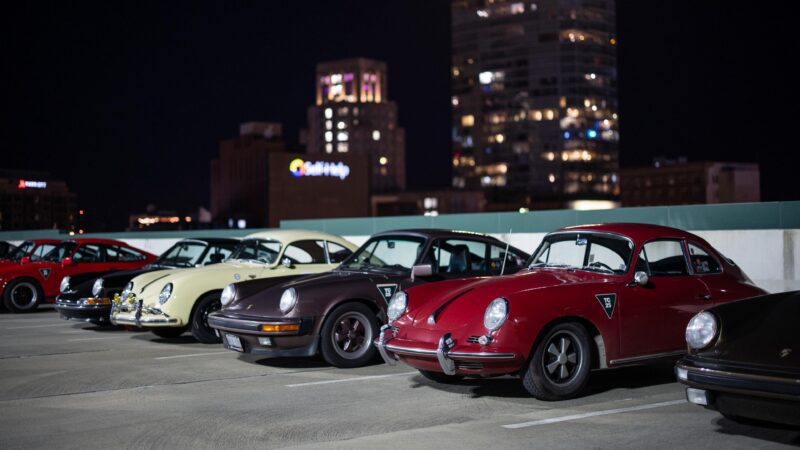 Several classic Porsche cars, including a Porsche 993, are parked in a row on a rooftop parking lot at night during Luftgekühlt 11 in Durham NC, with the city skyline glowing in the background.