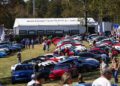 A large outdoor car show features dozens of parked Porsche vehicles, including 2025 models, with many people walking around and a white Porsche-branded tent in the background, celebrating IMSA WeatherTech SportsCar excitement.