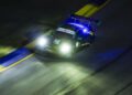 A Porsche race car with headlights on speeds along a dimly lit track at night during the IMSA WeatherTech SportsCar event, blue and yellow curbs visible at the road's edge.