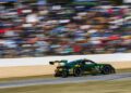A green and yellow Porsche race car speeds along the track at the 2025 IMSA WeatherTech SportsCar event, with a blurred crowd of spectators in the background.
