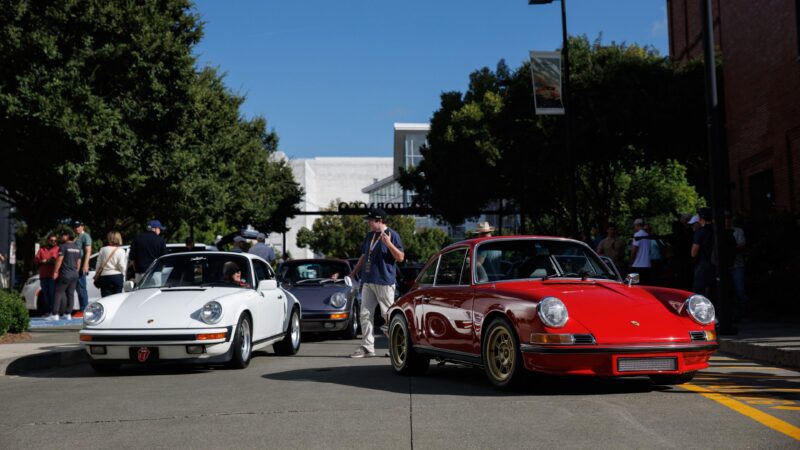 Three classic Porsche sports cars, including a stunning Porsche 993, are parked on a street as people look on at Luftgekühlt 11 in Durham NC under clear blue skies.