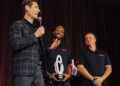 A man in a plaid blazer speaks into a microphone on stage while two men in black shirts, one holding a trophy, stand beside him and smile.