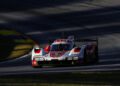 A red and white Porsche race car with visible dirt on the front drives on a sunlit section of the IMSA WeatherTech SportsCar racetrack, casting shadows in the background, embodying the thrill of 2025 motorsport.