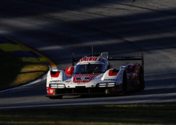 A red and white Porsche race car with visible dirt on the front drives on a sunlit section of the IMSA WeatherTech SportsCar racetrack, casting shadows in the background, embodying the thrill of 2025 motorsport.