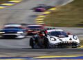 A white SportsCar with a shark mouth design leads a group of cars on the racetrack during an IMSA WeatherTech event, with motion blur highlighting the high-speed action.