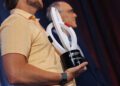 A man in a yellow shirt holds a sculptural trophy while standing next to another man in a suit on a stage.