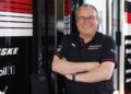 A smiling man wearing glasses and a Team Penske polo shirt stands with arms crossed in front of a Penske-branded garage, ready for the 2025 IMSA WeatherTech SportsCar season with Porsche.