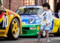 A young boy wearing a Mickey Mouse sweater points at a yellow Porsche 993, with another colorful race car parked behind him on a brick street in Durham NC during Luftgekühlt 11.