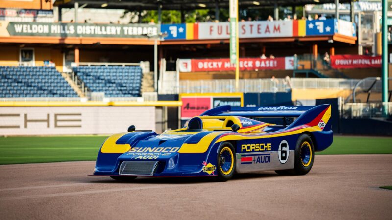 A blue and yellow vintage Porsche race car with Sunoco branding, reminiscent of the classic Porsche 993, is parked on a baseball field near empty stands at Luftgekühlt 11 in Durham NC.