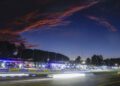 Porsche race cars speed by a brightly lit pit lane at dusk during the 2025 IMSA WeatherTech SportsCar event, with a dramatic, colorful sky and dark trees in the background.