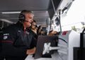 A man wearing a headset and Porsche team uniform monitors controls and screens at the 2025 IMSA WeatherTech SportsCar pit wall, with other crew members in the background.
