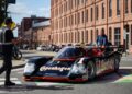 A person directs traffic as a man drives a vintage Porsche 993 with Copenhagen livery during Luftgekühlt 11 on a city street lined with brick buildings in Durham NC.