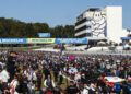 A large crowd gathers on the racetrack grid next to parked Porsche race cars, with Michelin banners and the Michelin Man visible in the background during the 2025 IMSA WeatherTech SportsCar event.