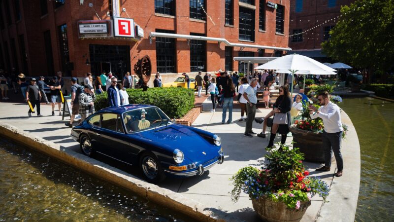 A blue Porsche 993 is parked by a pond at an outdoor event in Durham NC, in front of a brick building, with people socializing and gathering around during Luftgekühlt 11.