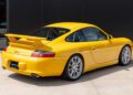 A yellow Porsche 911 GT3 sports car, a National Winner of the Porsche Classic Restoration 2025, is parked on a concrete surface in front of a black wall.